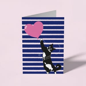 May include: A folded greeting card featuring a black and white cat with a white chest and paws, reaching up to hold a pink heart-shaped balloon. The background consists of horizontal dark blue and light pink stripes across the entire card surface.