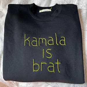 May include: Black sweatshirt with the words "Kamala is brat" embroidered in yellow thread. The sweatshirt is folded neatly on a floral patterned surface. A small tag is visible near the neckline.