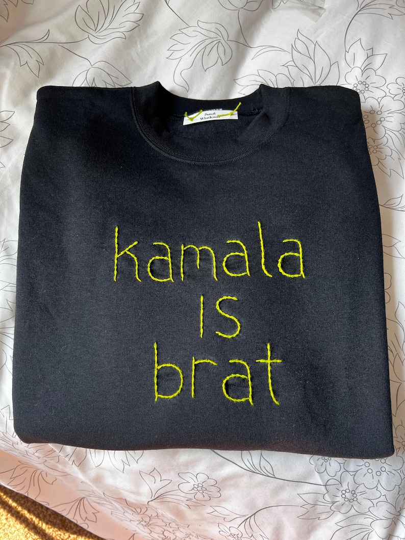 May include: Black sweatshirt with the words "Kamala is brat" embroidered in yellow thread. The sweatshirt is folded neatly on a floral patterned surface. A small tag is visible near the neckline.