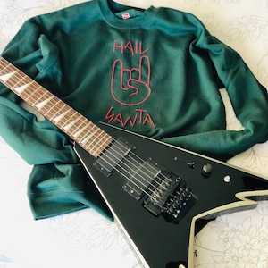May include: A dark green sweatshirt with the embroidered text "HAIL SANTA" and a rock-and-roll hand gesture in red. A black electric guitar with a V-shaped body rests on the sweatshirt.