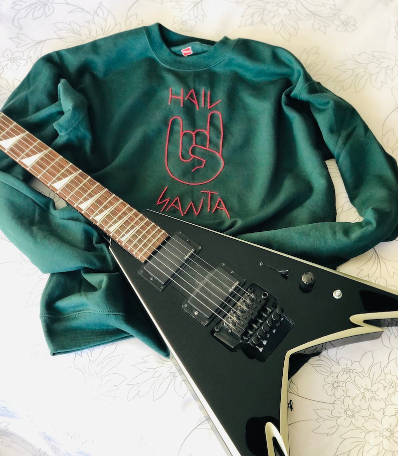May include: A dark green sweatshirt with the embroidered text "HAIL SANTA" and a rock-and-roll hand gesture in red. A black electric guitar with a V-shaped body rests on the sweatshirt.