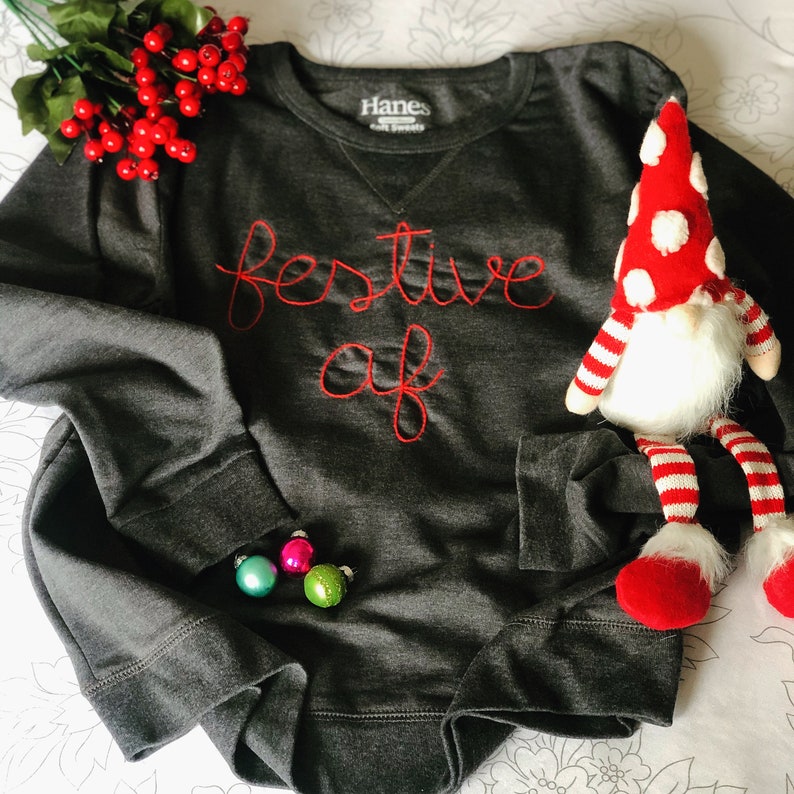 May include: Dark gray sweatshirt with the embroidered text "festive af" in red. The sweatshirt is next to a Christmas gnome and small ornaments. The brand name "Hanes" is visible on the tag.