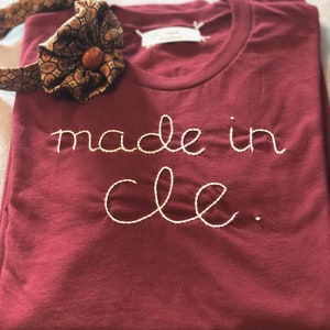 May include: Burgundy t-shirt with the embroidered text "made in cle." A patterned bow tie with a brown button is placed on the shirt. The shirt is folded neatly.