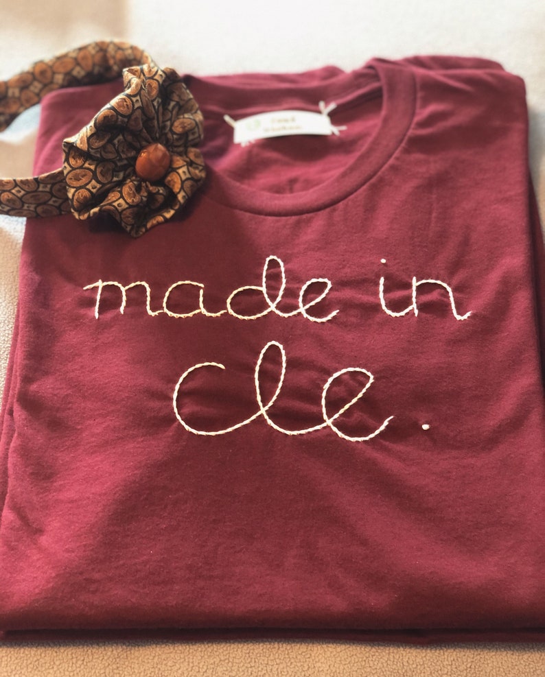 May include: Burgundy t-shirt with the embroidered text "made in cle." A patterned bow tie with a brown button is placed on the shirt. The shirt is folded neatly.