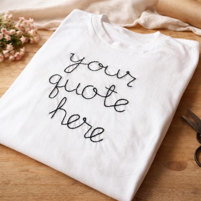 May include: A white t-shirt with the embroidered text "your quote here" in black cursive script. The shirt is neatly folded, suggesting it's ready to wear. The design is simple and elegant, suitable for personalization.