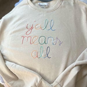 May include: A beige sweatshirt with the embroidered phrase "y'all means all" in rainbow-colored thread. The crew neck sweatshirt has long sleeves and a relaxed fit, perfect for casual wear.