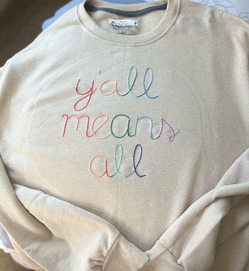 May include: A beige sweatshirt with the embroidered phrase "y'all means all" in rainbow-colored thread. The crew neck sweatshirt has long sleeves and a relaxed fit, perfect for casual wear.