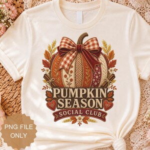 Pode incluir: Uma t-shirt cor de creme com um gráfico de uma abóbora decorada com um laço xadrez, espigas de trigo e paus de canela. O texto PUMPKIN SEASON SOCIAL CLUB está em letras castanho-escuras, rodeado por pequenos corações e detalhes florais.