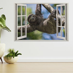 May include: A sloth hanging upside down from a tree branch, looking out of a window. The sloth is brown and has a white face. The window is white and has a white frame. The background is a blurry green and blue.