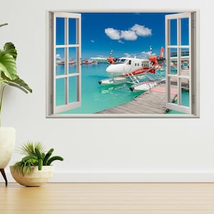 May include: A white seaplane with red accents is docked at a wooden pier over turquoise water. The seaplane is in front of a window with a view of a tropical island.