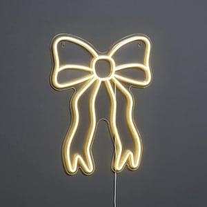 Op de afbeelding: Een neonlicht in de vorm van een strik, met een warme gele gloed. De omtrek van de strik is verlicht, met twee lange, vloeiende linten. Het licht is bevestigd aan een donkergrijze muur.