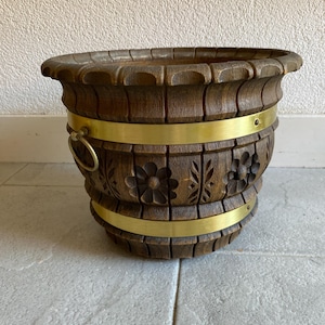 May include: A wooden planter with a carved floral design and brass bands. The dark brown planter has a decorative rim and a brass ring handle. The planter is sitting on a gray tiled surface.