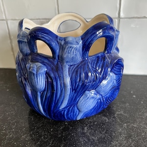 May include: A blue ceramic planter with a floral design. The planter features tulip-like shapes around the exterior and openings at the top. The interior is a light cream colour. The planter is approximately 15 cm in diameter.