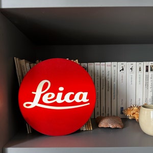 Puede incluir: Un letrero circular rojo con la palabra "Leica" en escritura blanca en una estantería con libros y una escultura blanca.