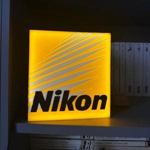 Może przedstawiać: Podświetlony znak w kolorze żółtym i białym z napisem "Nikon" czarnymi literami.