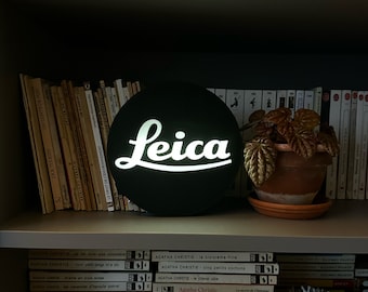 May include: A black and white sign with the word "Leica" in white letters on a black background. The sign is hanging on a shelf with books and a potted plant.