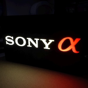 Puede incluir: Un letrero negro con las palabras "SONY α" en letras blancas y rojas. El "SONY" está en blanco y el "α" está en rojo.