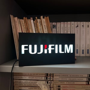 Może przedstawiać: Czarny znak z białymi literami, na którym widnieje napis "FUJIFILM", na białej półce z książkami ułożonymi po obu stronach.