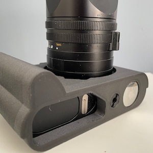 Pode incluir: Uma câmera preta com uma lente preta. A câmera tem uma capa protetora preta com uma alça. A lente tem o texto "MADE IN GERMANY" impresso.