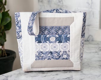 May include: A quilted tote bag with a patchwork design. The bag features a combination of white, blue, and gray fabrics. The main panel has floral and geometric patterns. The handles are made of the same blue floral fabric.