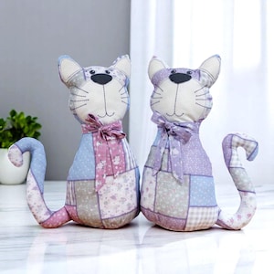 May include: Two cat-shaped doorstops made from patchwork fabric in pastel shades of pink, blue, and lavender. They feature embroidered faces, a bow tie, and a curled tail. The doorstops are designed to stand upright and are made from various fabric patterns.