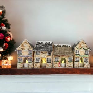 May include: Four miniature houses with lit candles inside, each with a different storefront sign: "The Haberdashery", "The Royal Oak", "Old Post Office", and "Tea Rooms".