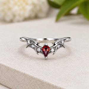 Puede incluir: Un anillo de tono plateado con una piedra preciosa roja central en forma de cometa, flanqueada por estructuras metálicas en forma de alas. Las alas están acentuadas con cuatro pequeñas piedras transparentes de corte redondo incrustadas.