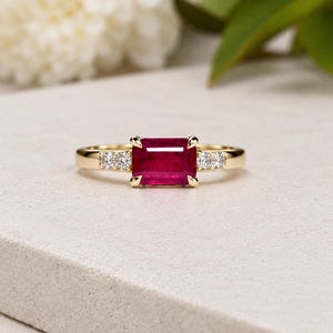 May include: A gold ring featuring a rectangular, deep red ruby gemstone set in the center, flanked by three small, clear, round-cut diamonds on each side of the band. The metal band is polished yellow gold with a smooth, minimalist design.