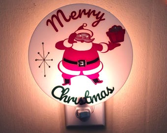 May include: A circular plug-in night light featuring a red and white graphic of Santa Claus holding a gift. The text Merry Christmas is printed in red and green script. A starburst icon is on the left. The light emits a warm glow from behind.