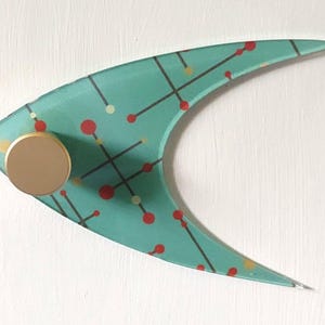 May include: A teal-colored, mid-century modern wall hook with a gold-colored knob. The hook has a unique boomerang shape and is decorated with a pattern of red and yellow dots and black lines. The hook is mounted on a white surface.