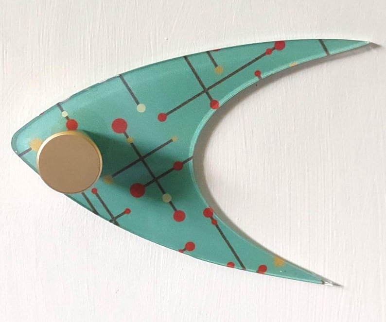May include: A teal-colored, mid-century modern wall hook with a gold-colored knob. The hook has a unique boomerang shape and is decorated with a pattern of red and yellow dots and black lines. The hook is mounted on a white surface.