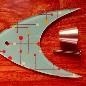 May include: A mid-century modern fish-shaped cabinet pull in aqua with red, yellow, and white dot and line design. Includes a silver-toned knob and screw. The hardware is set against a reddish-brown wooden surface.