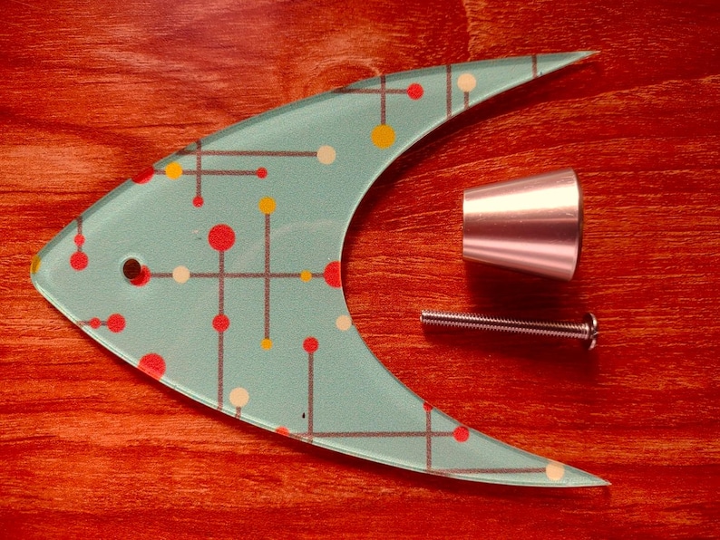 May include: A mid-century modern fish-shaped cabinet pull in aqua with red, yellow, and white dot and line design. Includes a silver-toned knob and screw. The hardware is set against a reddish-brown wooden surface.