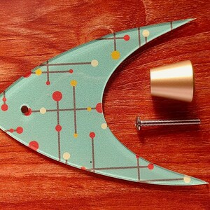 May include: A decorative fish-shaped glass cabinet pull with a light blue background and a pattern of red, yellow, and white dots and lines. Includes a gold-colored knob and a silver screw. The items are on a reddish-brown wooden surface.