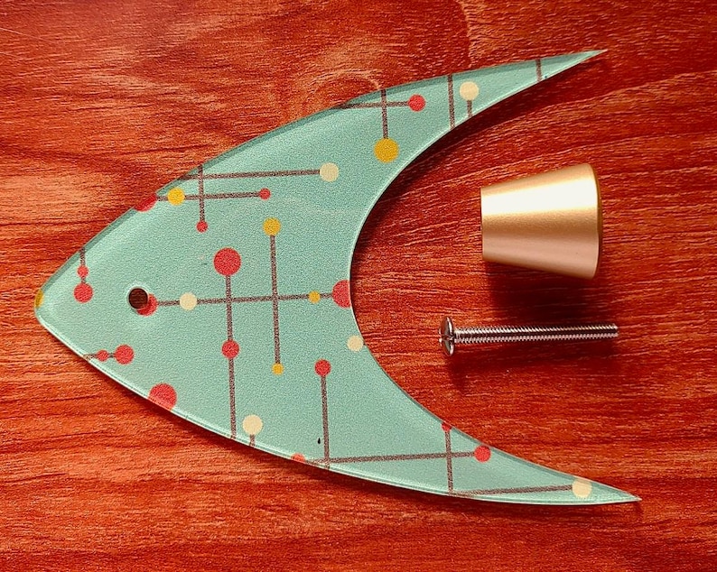 May include: A decorative fish-shaped glass cabinet pull with a light blue background and a pattern of red, yellow, and white dots and lines. Includes a gold-colored knob and a silver screw. The items are on a reddish-brown wooden surface.