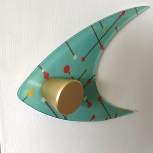 May include: A mid-century modern style cabinet pull with a teal, curved design. The pull features a gold-colored knob and a pattern of red, yellow, and white dots connected by black lines. The cabinet pull is mounted on a white surface.