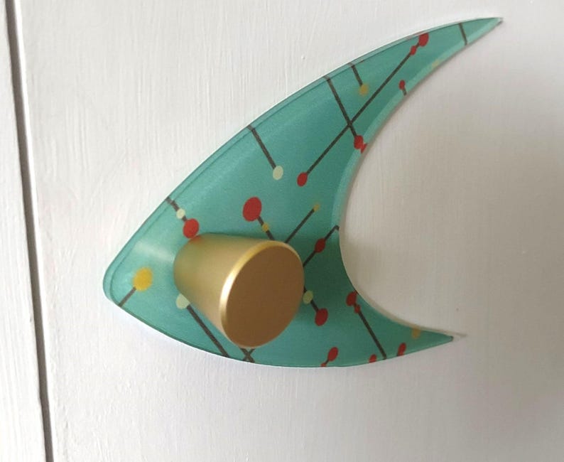 May include: A mid-century modern style cabinet pull with a teal, curved design. The pull features a gold-colored knob and a pattern of red, yellow, and white dots connected by black lines. The cabinet pull is mounted on a white surface.