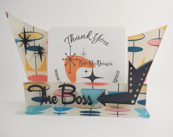 May include: A clear acrylic desk sign featuring a colorful pattern of teal, pink, and yellow shapes with black starbursts. The sign displays the text The Boss in black script and includes a black arrow pointing right, with a card reading Thank You RetroModDesigns.