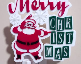 May include: A decorative night light featuring a white acrylic cutout with the red text Merry and green text CHRIST MAS. A red and white Santa Claus figure is centered, pointing toward the text, accented by small white snowflake shapes.