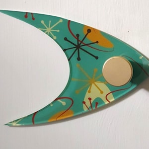 Peut inclure: Un crochet mural de style moderne du milieu du siècle, de couleur turquoise, avec un bouton doré. Le crochet a une forme incurvée unique et est décoré d'un motif rétro d'éclats d'étoiles et de tourbillons jaunes, rouges et noirs.