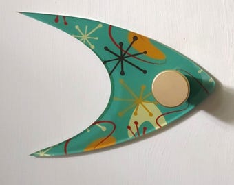 May include: A teal, mid-century modern style wall hook with a gold-colored knob. The hook has a unique, curved shape and is decorated with a retro atomic pattern in yellow, red, and black. It is mounted on a white surface.