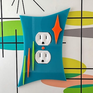 Puede incluir: Una tapa de tomacorriente azul turquesa con dos tomas de corriente estándar. La tapa tiene un diseño moderno con formas geométricas naranja, verde y verde lima.