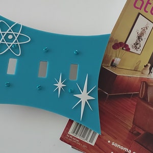 May include: A turquoise blue double light switch cover with a white atomic symbol and two white starburst designs. The cover is made of a smooth, glossy material and has a slightly curved shape.