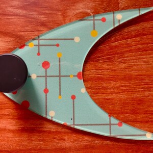 May include: A mid-century modern style glass serving piece with a teal background and a pattern of red, yellow, and white dots and lines. A black, round handle is attached to the side. The piece is on a reddish-brown wooden surface.