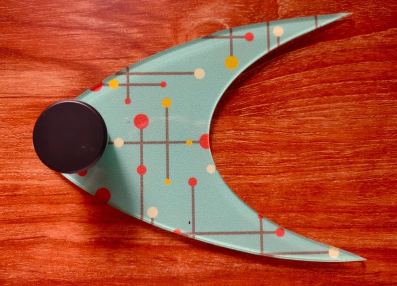 May include: A mid-century modern style glass serving piece with a teal background and a pattern of red, yellow, and white dots and lines. A black, round handle is attached to the side. The piece is on a reddish-brown wooden surface.