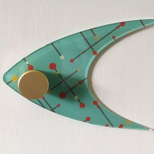 May include: A teal, boomerang-shaped wall hook with a mid-century modern design. The hook features a gold-colored knob and a pattern of red, yellow, and black dots and lines. The hook is mounted on a white surface.