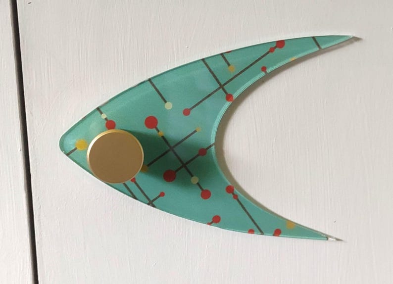 May include: A teal, boomerang-shaped wall hook with a mid-century modern design. The hook features a gold-colored knob and a pattern of red, yellow, and black dots and lines. The hook is mounted on a white surface.
