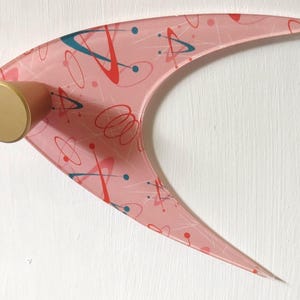 Peut inclure: Un élément de décoration murale rose en forme de boomerang de style moderne du milieu du siècle, avec une fixation cylindrique dorée. Le boomerang présente un motif de formes abstraites rouges, bleues et blanches. La décoration est fixée sur un mur blanc.