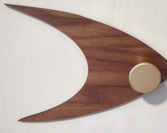 May include: A mid-century modern wall decor piece. The wooden art has a dark brown finish and a unique boomerang shape. A gold-colored, circular accent is attached to the wood. The art is mounted on a white wall.