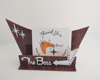 May include: A dark wood-grain desk sign holder featuring a boomerang shape with white star accents. The front displays the white text The Boss next to a white arrow. A card inside reads Thank You with a boomerang graphic and RetroModDesigns.